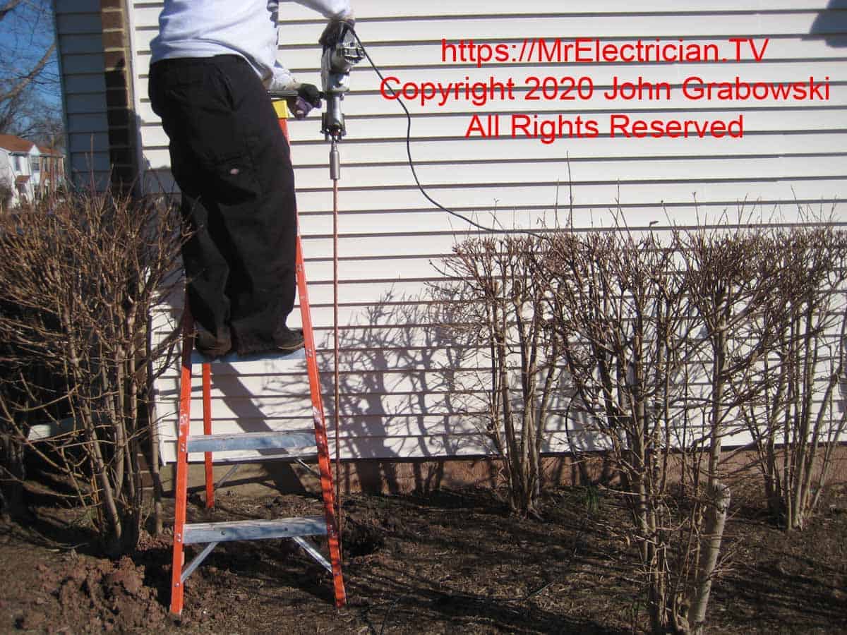Man on a ladder using a rotary hammer to drive a ground rod into the ground
