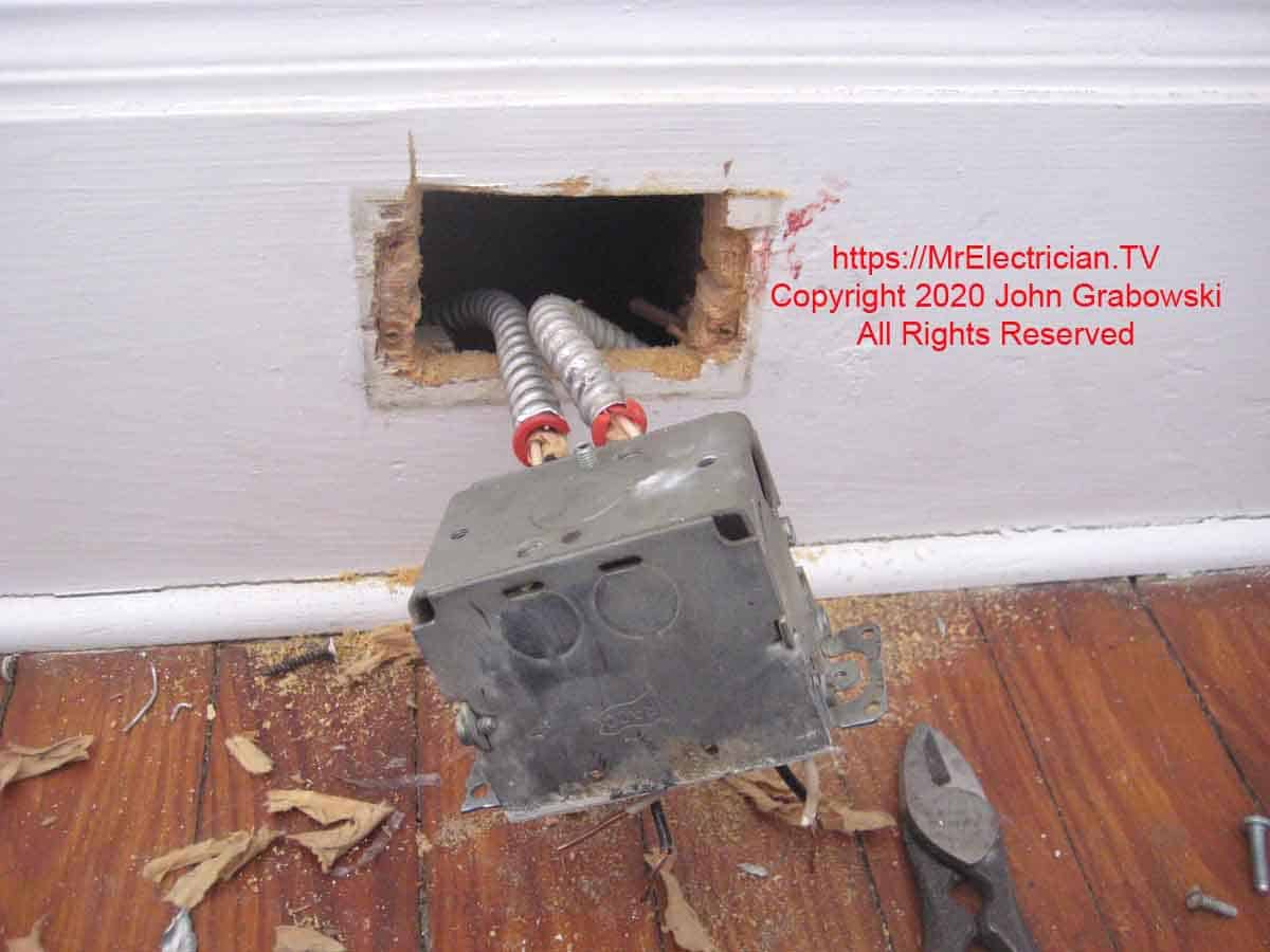 Armored cables in the wood baseboard with red anti-short bushings, entering the bottom of a metal outlet box before its insertion into the wall. With this type of metal outlet box the cable clamp can be used on all four sides. However, each clamp used requires a deduction of one wire allowed in the box. Wiring devices such as outlets require two wire deductions.