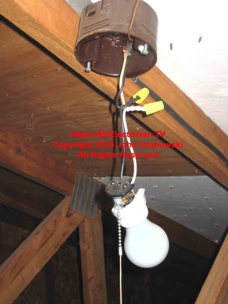 An old broken light bulb socket with a pull chain hanging by wires from a brown plastic electrical box in an attic.
