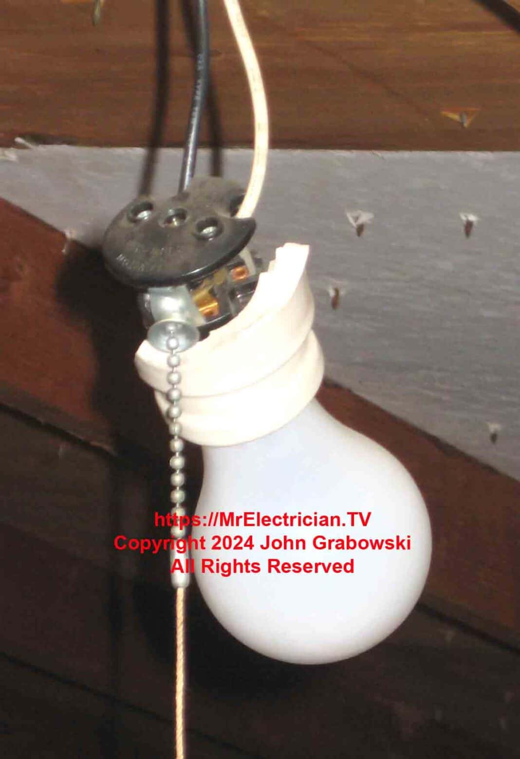A close shot of a broken light bulb socket with a pull chain hanging by two electrical wires in an attic.