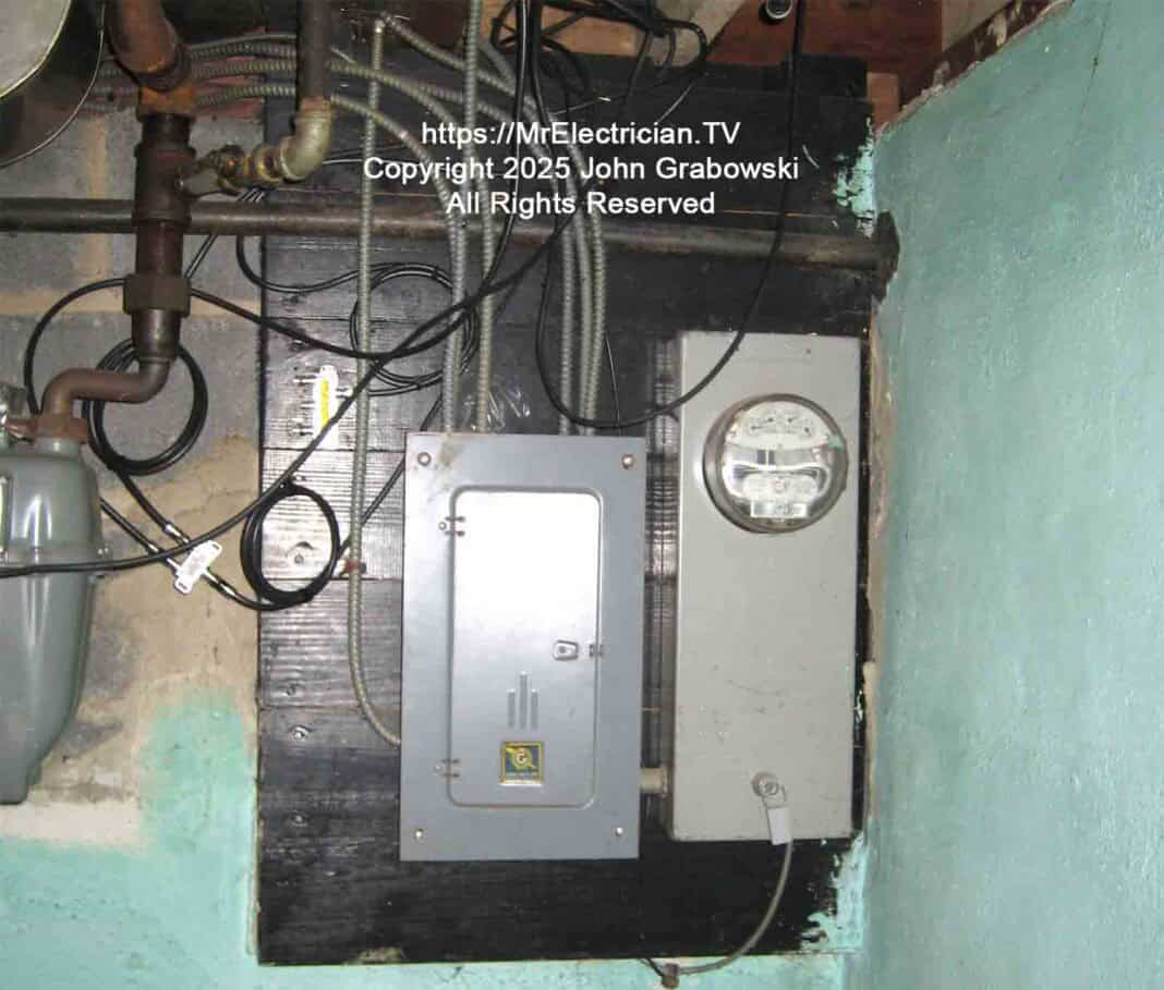 An older electric meter and fuse box located inside a basement adjacent to the foundation wall. The armored grounding electrode conductor is coming out of the bottom of the meter socket.