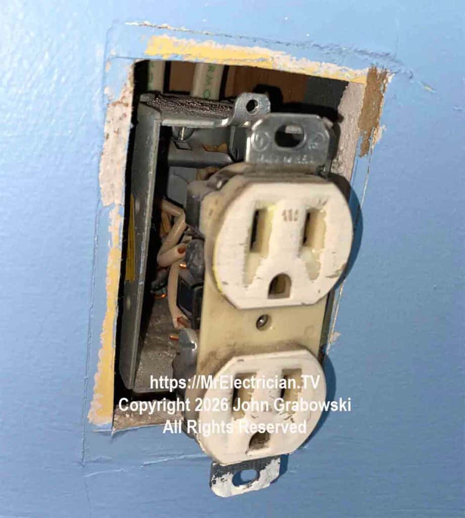 An electrical receptacle outlet with the mounting screws removed so the outlet is only held in place by the wires connected to it