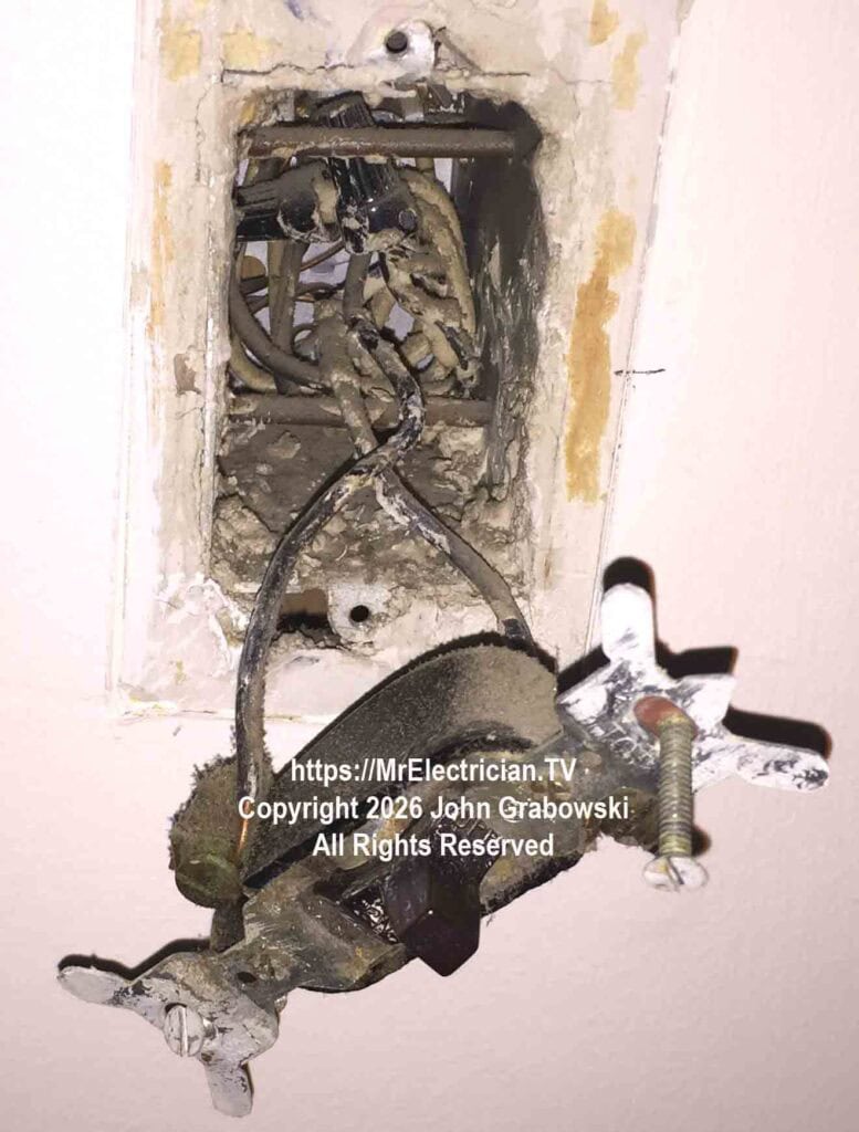 An old electrical wall switch hanging by two black wires coming from the metal switch box inside the wall, which has a neutral wire. There are two 16D common nails passing through the upper and lower inside of the box to attach the switch box to the wood wall stud.