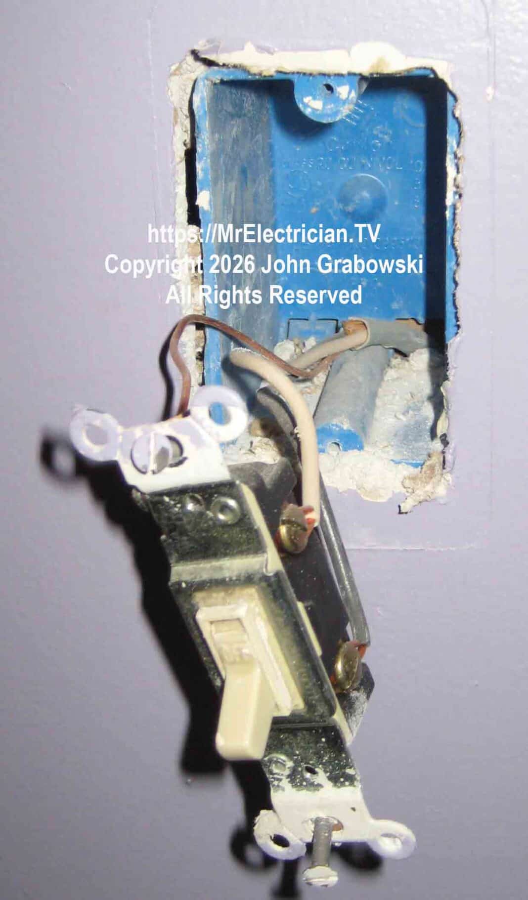 An ivory colored electrical wall switch hanging from a blue plastic switch box by a white wire, a black wire, and a bare copper wire. There is no neutral available here.